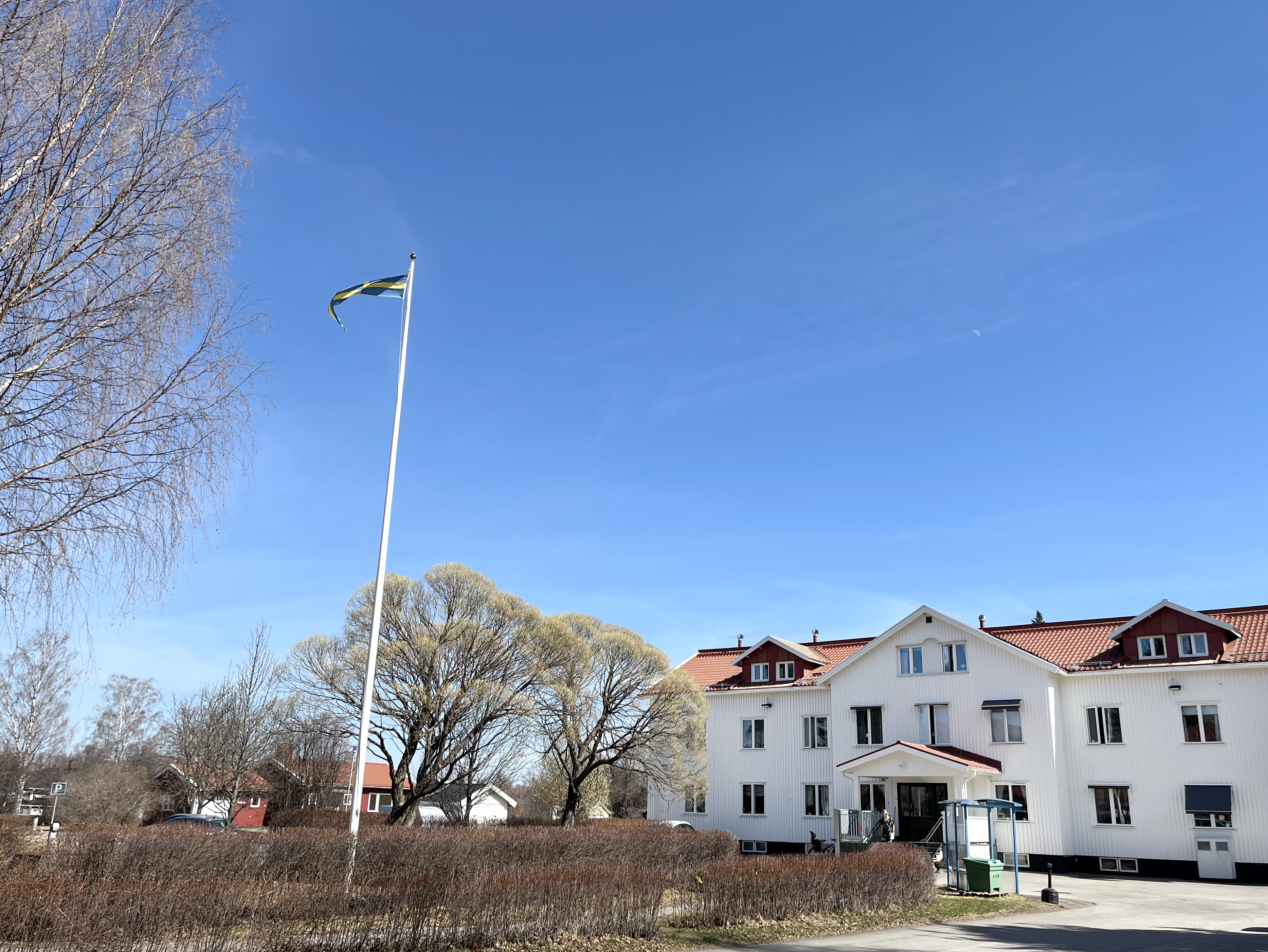 Bilden visar ett stort vitt hus. Framför huset syns en flaggstång och en häckplantering.