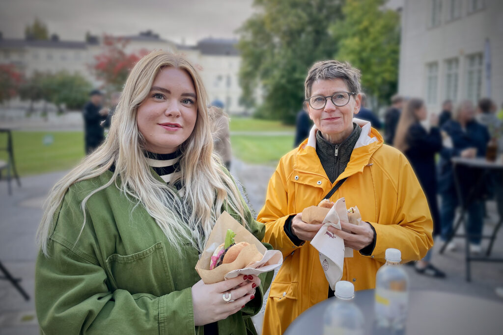 Bilden visar två kvinnor som tittar in i kameran. I bakgrunden syns människor mingla.