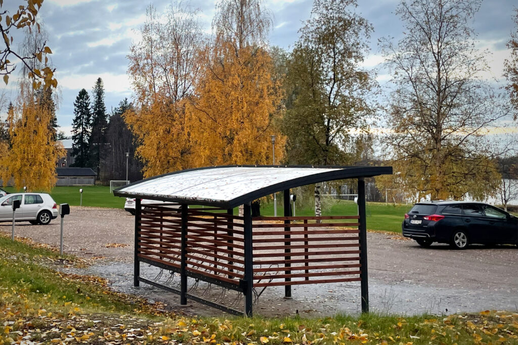 Bilden visar en cykelparkering i trä och stål. I bakgrunden syns höstlöv.