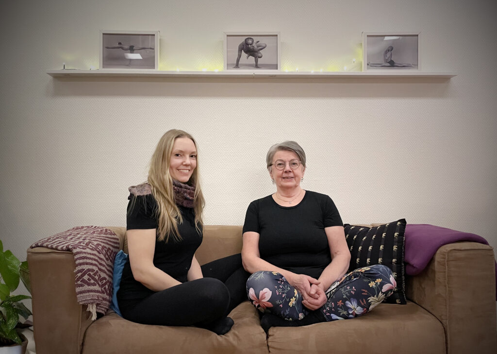 Två kvinnor sitter i en soffa. Båda tittar in i kameran och ler. Ovanför soffan hänger en hylla.