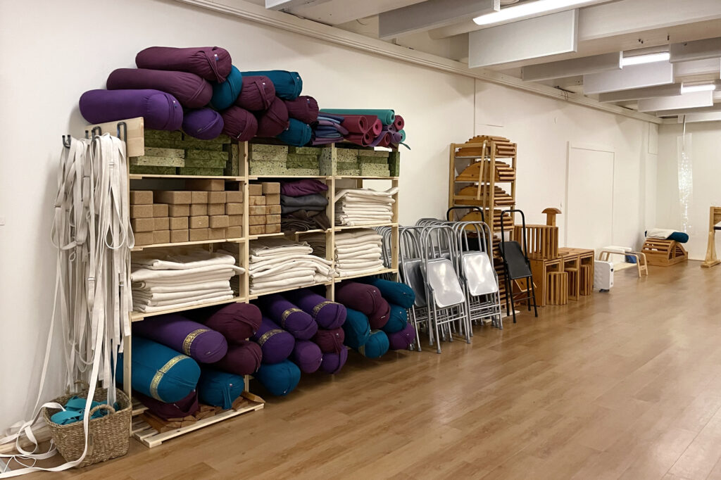 En hylla med yogatillbehör som mattor, klossar, rep och filtar står mot en vägg.