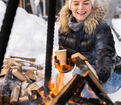 Young and beautiful woman wearing down jacket warming up by the fire pit during cold winter day